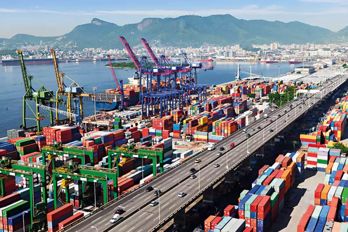 Panoramic view of an industrial port with cranes, multicolored cargo containers, an elevated highway, and a city in the background next to mountains Panoramic view of an industrial port with cranes, multicolored cargo containers, an elevated highway, and a city in the background next to mountains