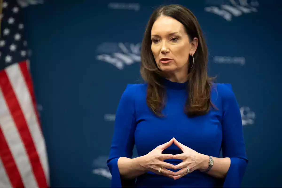 Brown-haired woman in a blue dress speaking in front of a blue background with a United States flag to the side Brown-haired woman in a blue dress speaking in front of a blue background with a United States flag to the side