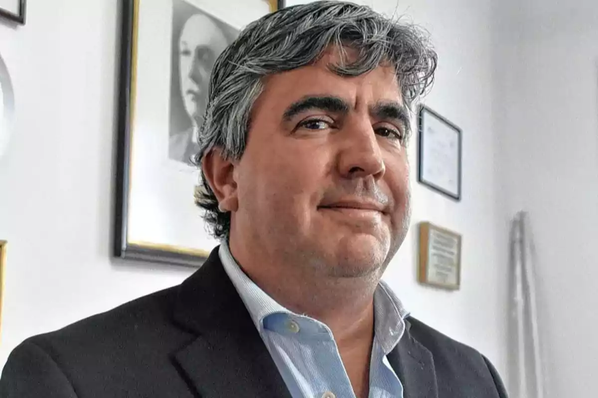 Hombre de cabello canoso y traje oscuro posando en una oficina con cuadros en la pared