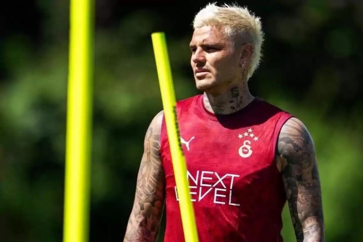 Futbolista rubio con tatuajes usando camiseta sin mangas roja del Galatasaray durante un entrenamiento al aire libre con postes amarillos en primer plano.