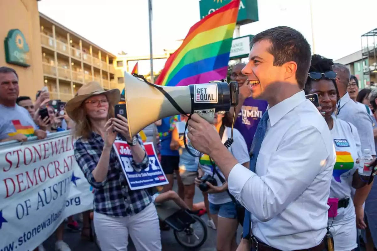 A smiling man in a jersey and tie uses a megaphone while participating in an LGBTQ+ pride march surrounded by people with rainbow flags and banners.