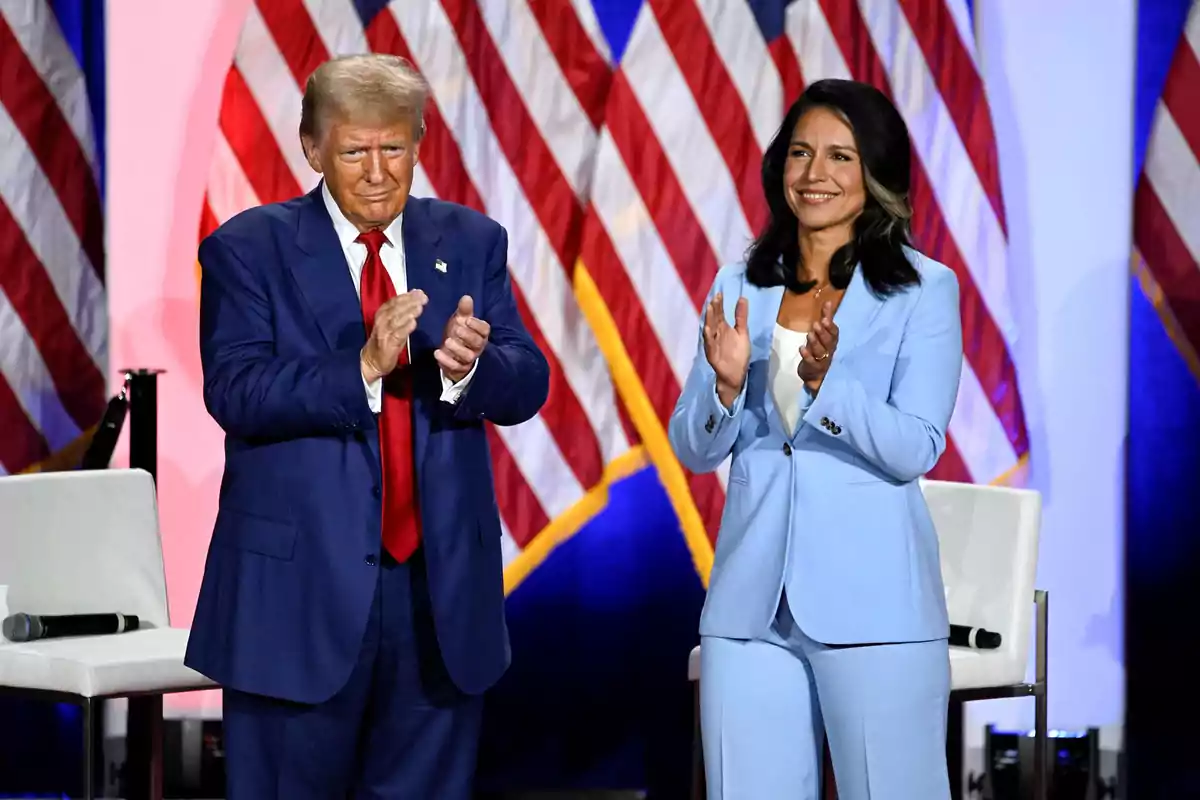 Dos personas de pie aplaudiendo en un escenario con varias banderas de Estados Unidos al fondo Dos personas de pie aplaudiendo en un escenario con varias banderas de Estados Unidos al fondo