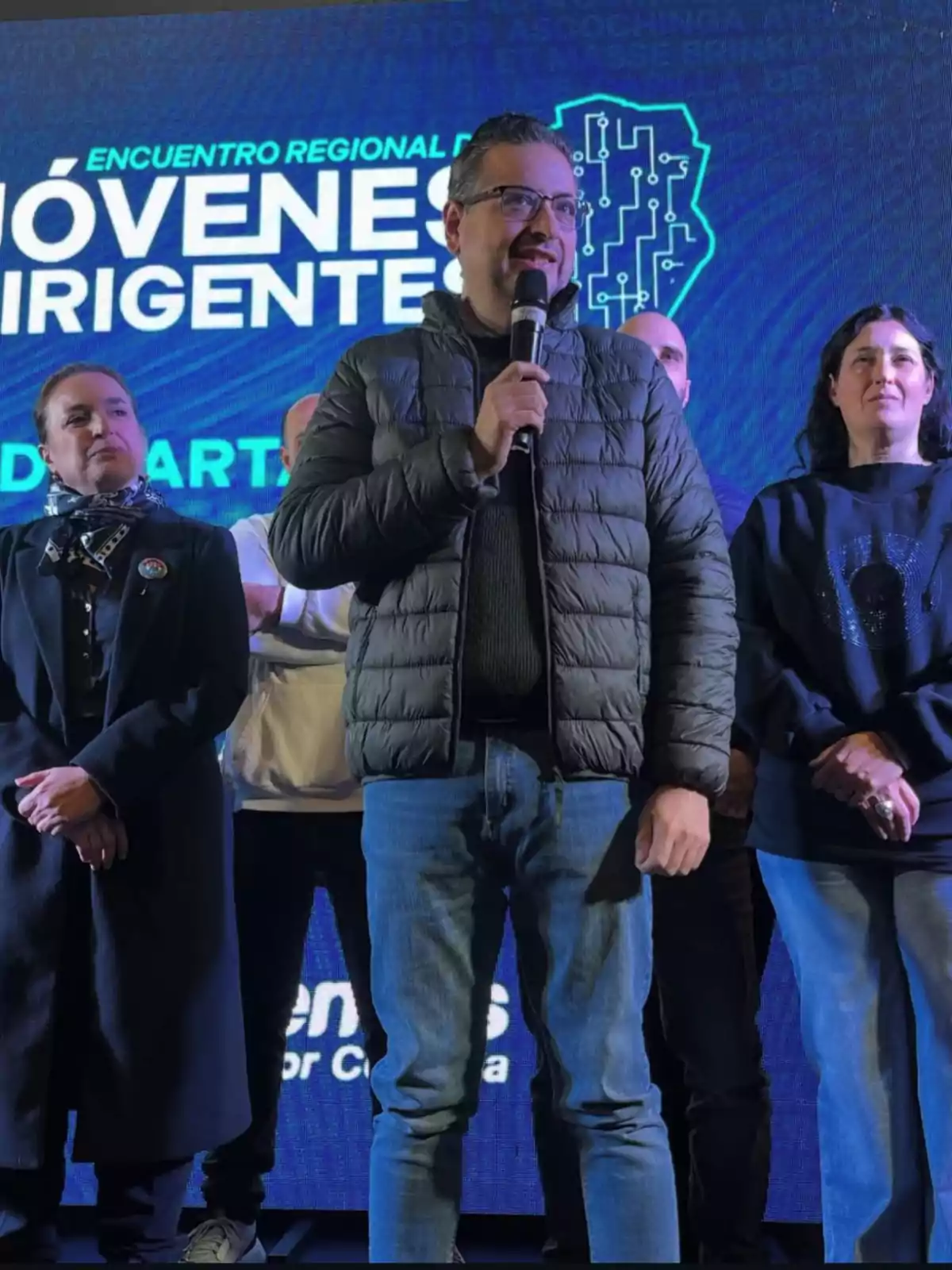 A man wearing a jacket and glasses holds a microphone while speaking on a stage alongside other people, with a blue background displaying an event for young leaders. A man wearing a jacket and glasses holds a microphone while speaking on a stage alongside other people, with a blue background displaying an event for young leaders.