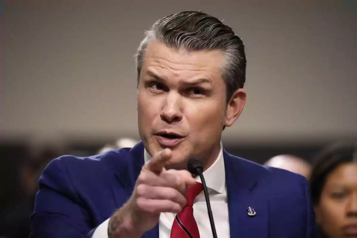 Short-haired, gray-haired man in a blue suit and red tie pointing with his finger while speaking in front of a microphone