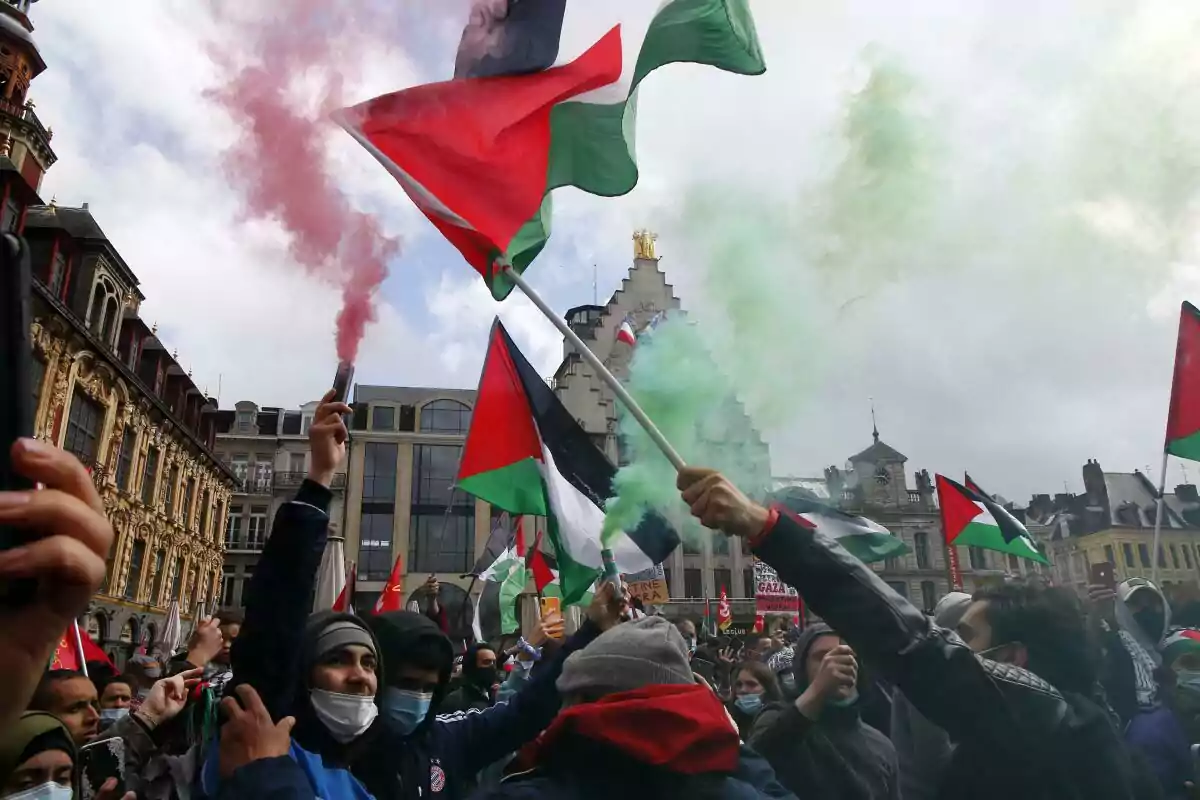 Demonstration in a square with people waving Palestinian flags and red and green smoke among old buildings Demonstration in a square with people waving Palestinian flags and red and green smoke among old buildings