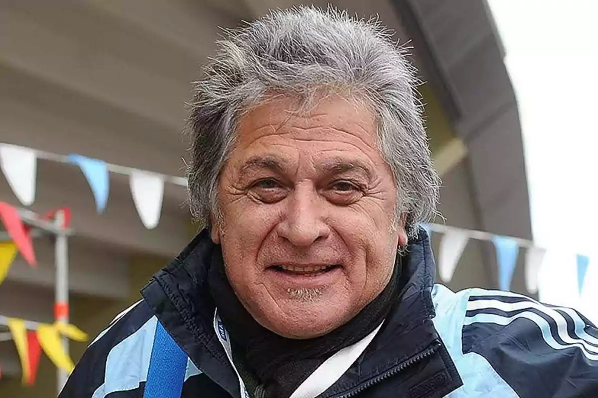 Hombre de cabello canoso y sonrisa vistiendo una chaqueta deportiva azul con banderines de colores al fondo