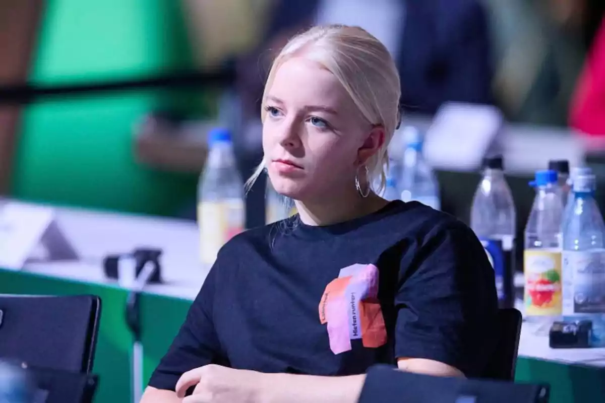 Joven de cabello rubio y camiseta negra sentada en una mesa con botellas de agua al fondo en un ambiente de conferencia Joven de cabello rubio y camiseta negra sentada en una mesa con botellas de agua al fondo en un ambiente de conferencia