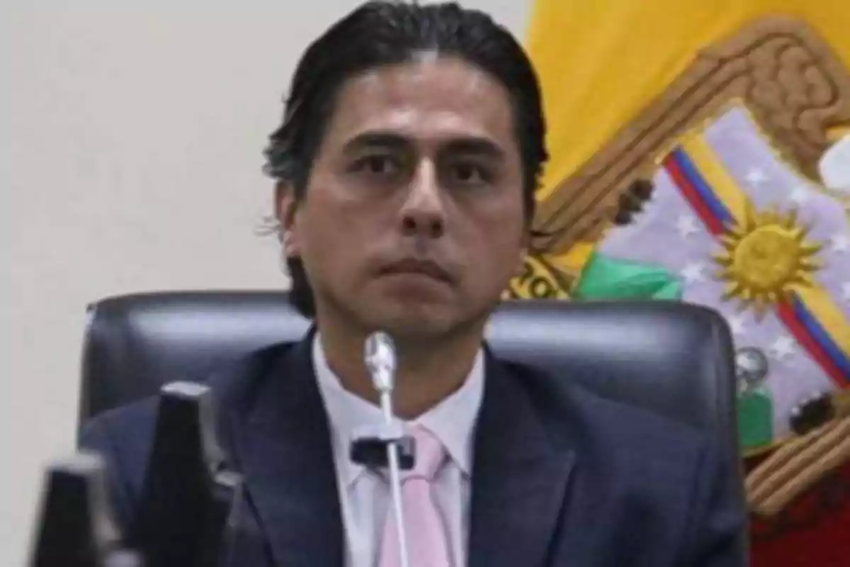 Dark-haired man in a formal suit sitting in front of a microphone with a flag in the background