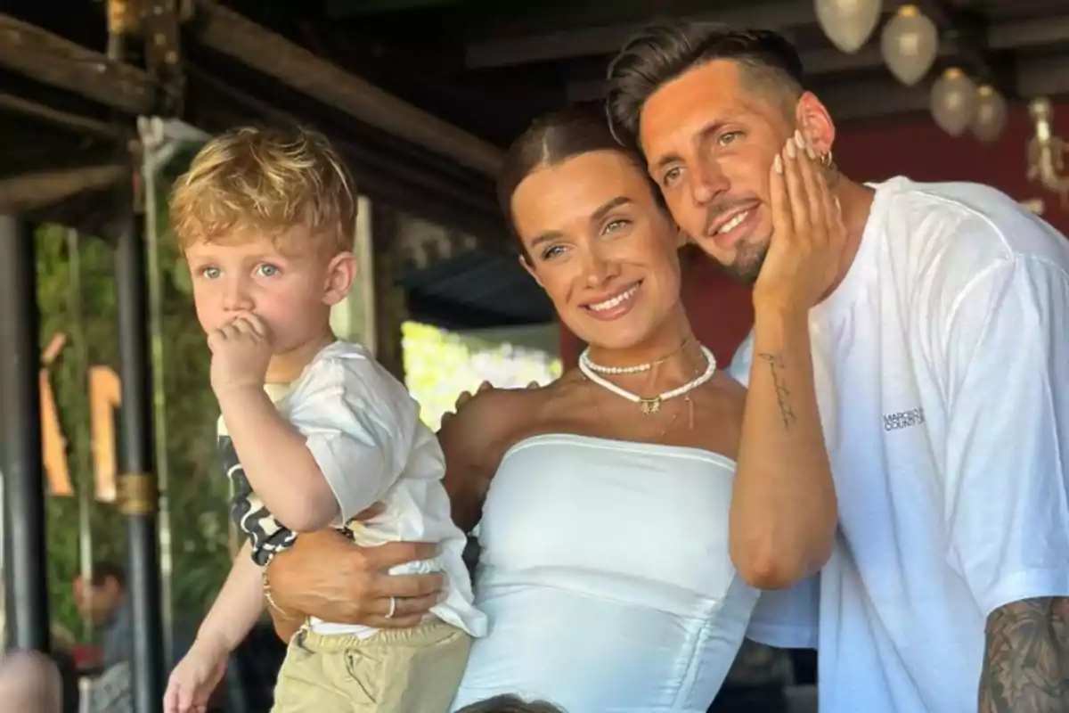 A family smiles and poses together outdoors; the woman is holding a small child while the man affectionately leans in.
