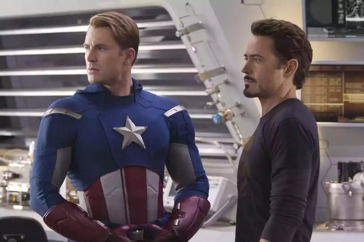Dos personajes de una película de superhéroes están de pie en lo que parece ser un laboratorio tecnológico uno lleva un traje azul con una estrella plateada en el pecho y el otro viste una camiseta oscura Dos personajes de una película de superhéroes están de pie en lo que parece ser un laboratorio tecnológico uno lleva un traje azul con una estrella plateada en el pecho y el otro viste una camiseta oscura