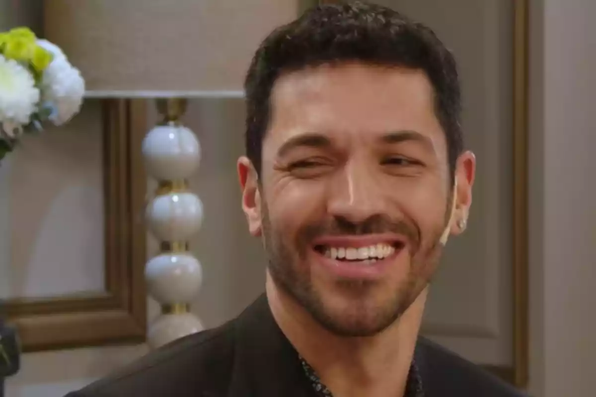 Short-haired, bearded man smiling while sitting in a room decorated with flowers and a lamp in the background. Short-haired, bearded man smiling while sitting in a room decorated with flowers and a lamp in the background.