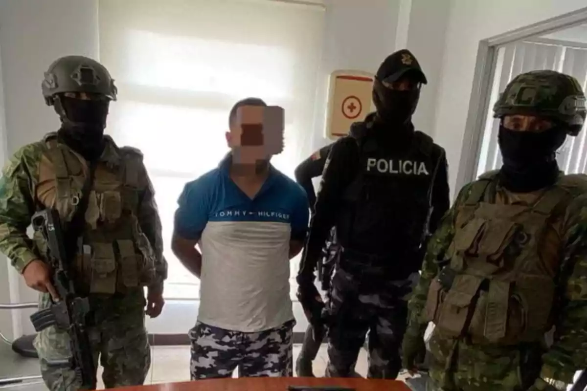 Tres hombres con uniforme militar y policial, armados y encapuchados, escoltan a un hombre con el rostro pixelado y las manos hacia atrás en una habitación iluminada.
