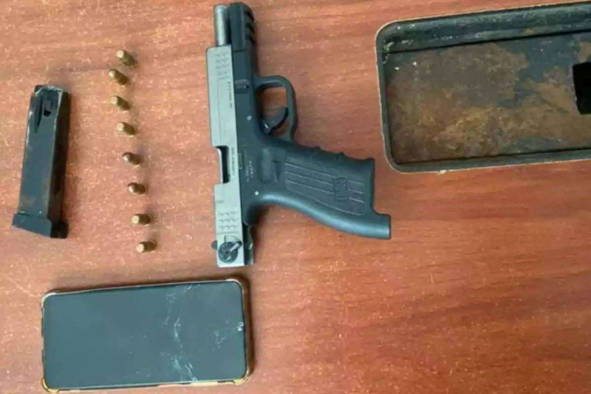 Pistola negra junto a un cargador, varias balas y un teléfono móvil sobre una mesa de madera Pistola negra junto a un cargador, varias balas y un teléfono móvil sobre una mesa de madera