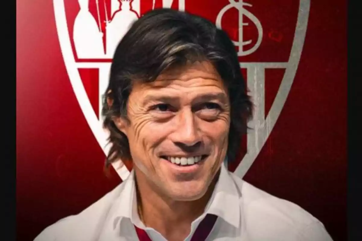 Hombre de cabello castaño sonriente con camisa blanca frente a un escudo rojo y blanco de un equipo de fútbol Hombre de cabello castaño sonriente con camisa blanca frente a un escudo rojo y blanco de un equipo de fútbol