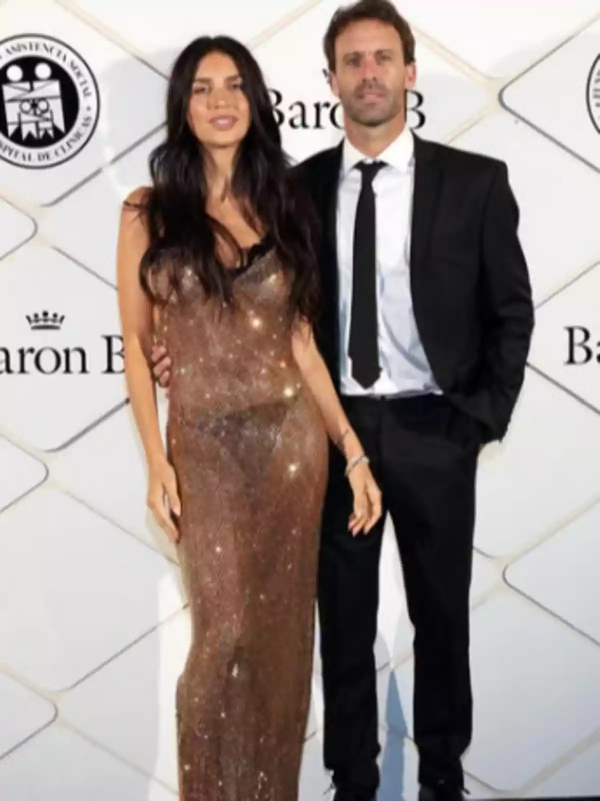 Una mujer con vestido dorado brillante y un hombre con traje negro posan juntos en un evento elegante frente a un fondo con logotipos.