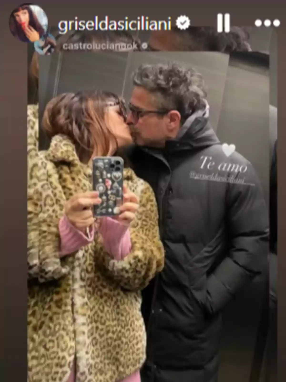 Pareja besándose frente a un espejo, ella lleva un abrigo de estampado animal y él un abrigo negro, ambos se ven reflejados mientras ella sostiene el teléfono para tomar la foto