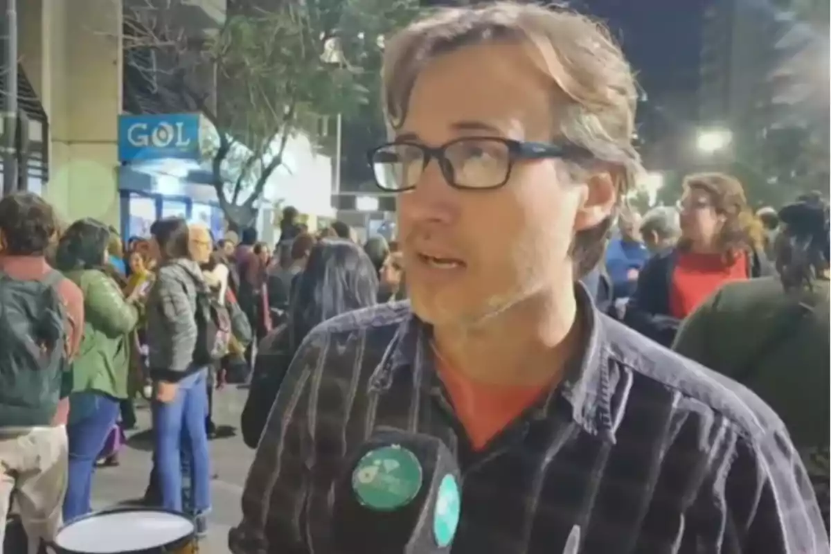 Hombre con gafas y camisa a cuadros hablando frente a un micrófono en una calle llena de personas durante la noche