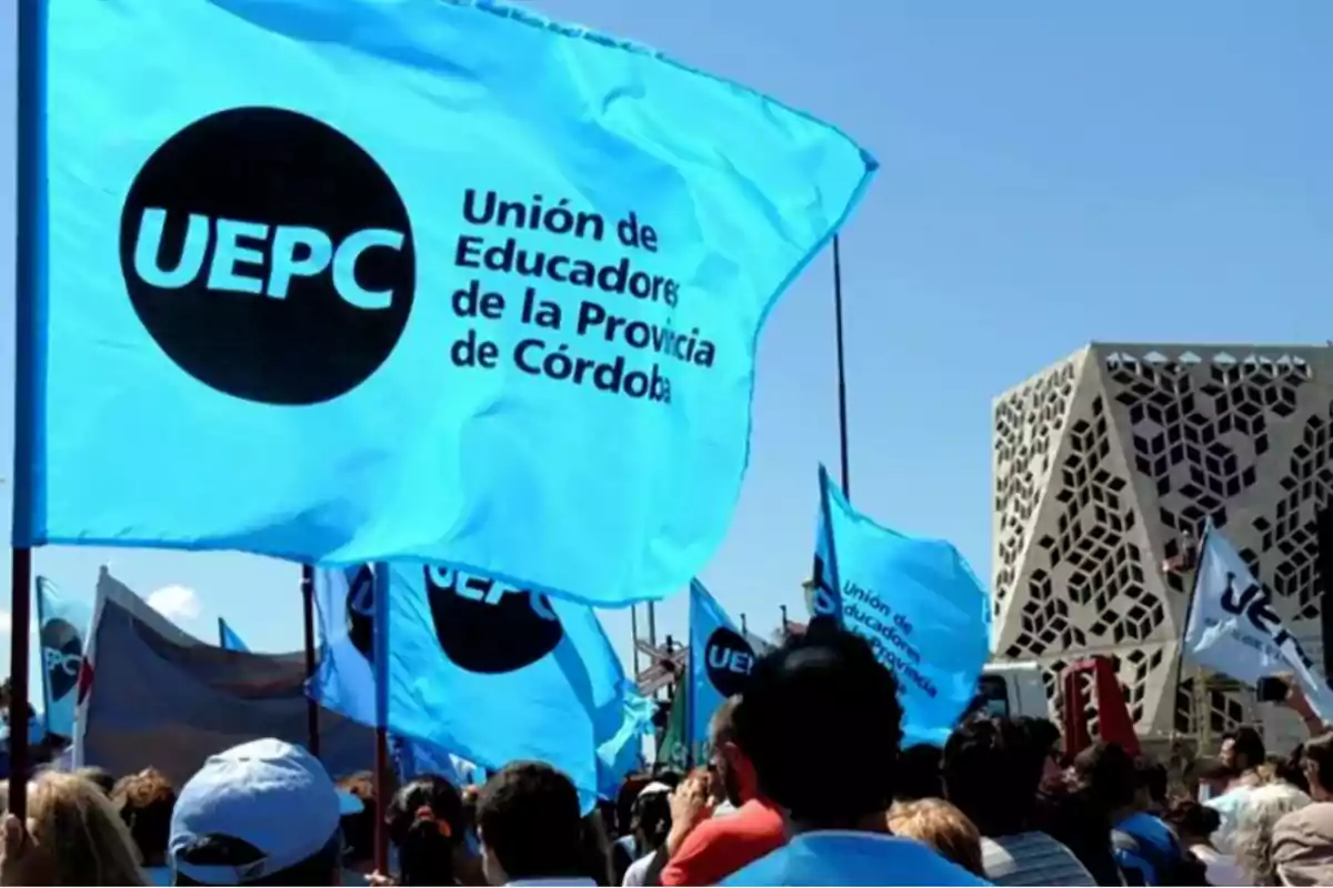 Manifestación de personas con banderas celestes de la Unión de Educadores de la Provincia de Córdoba frente a un edificio moderno