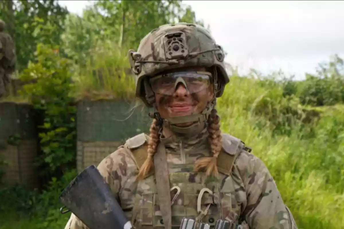 Female soldier in camouflage uniform and military helmet smiling in an outdoor setting surrounded by vegetation