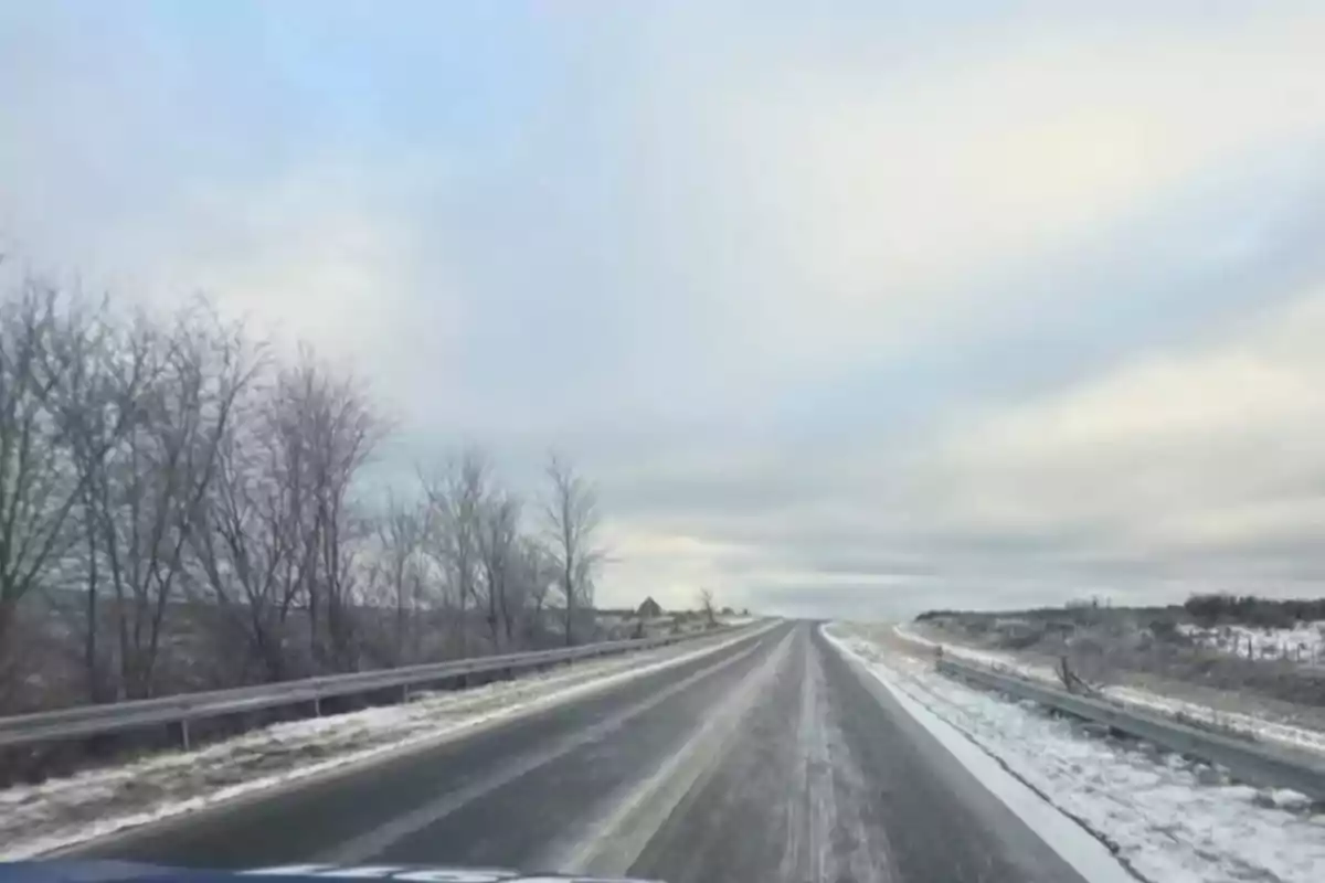Carretera cubierta parcialmente de hielo y nieve con árboles sin hojas a la izquierda y cielo nublado