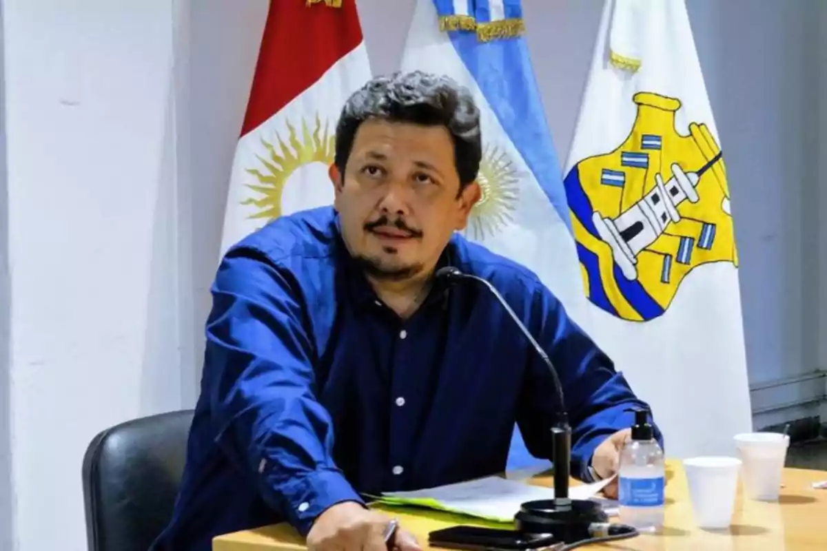Hombre de cabello corto y barba sentado en una mesa con micrófono y papeles, al fondo hay banderas argentinas y una botella de agua sobre la mesa