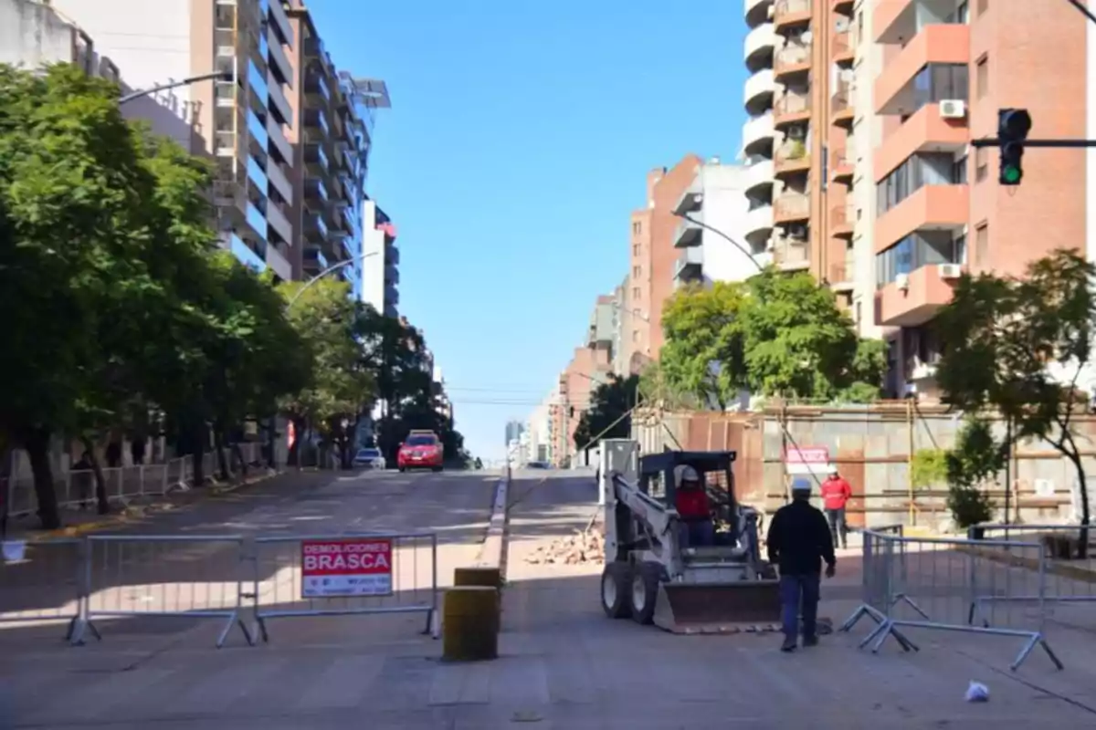 Calle urbana cerrada por obras con maquinaria y trabajadores, rodeada de edificios y árboles