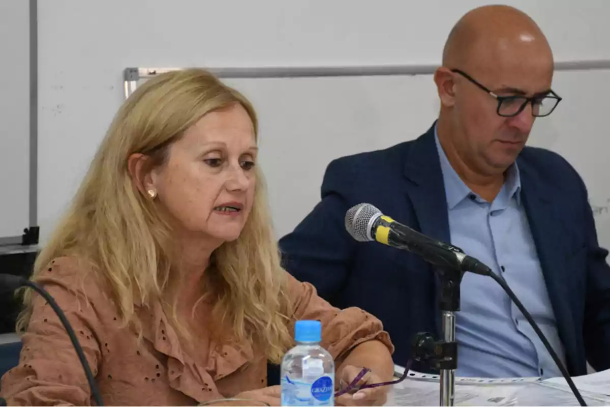 Mujer rubia hablando frente a un micrófono mientras un hombre con gafas revisa documentos a su lado en una mesa de trabajo