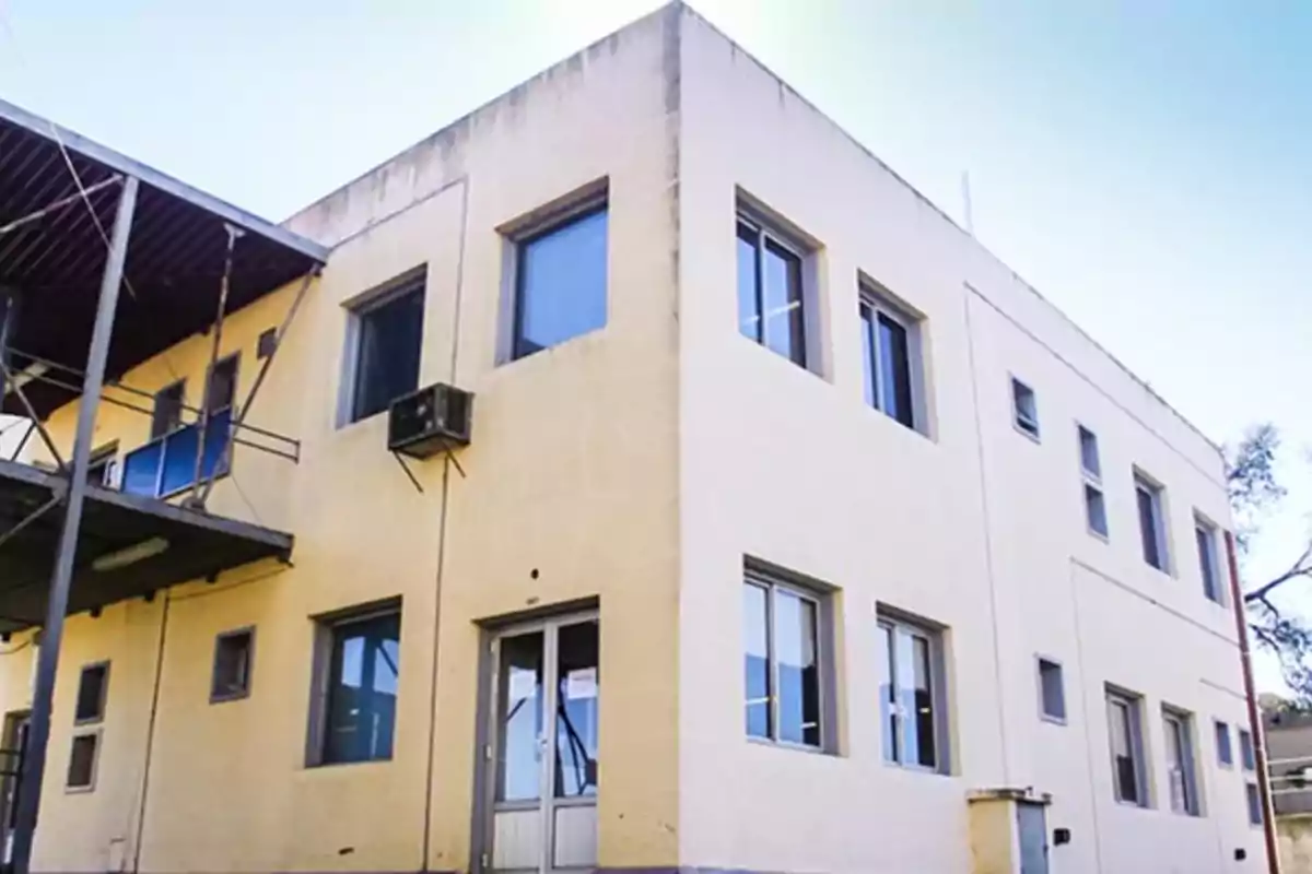 Edificio de dos pisos con paredes color beige y varias ventanas rectangulares, visto desde una esquina exterior en un día soleado