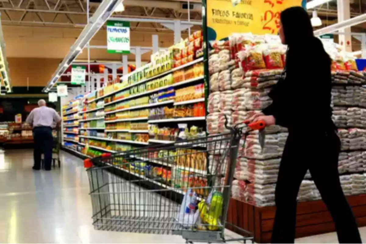 Persona empujando un carrito de compras en el pasillo de un supermercado con estantes llenos de productos y bolsas apiladas