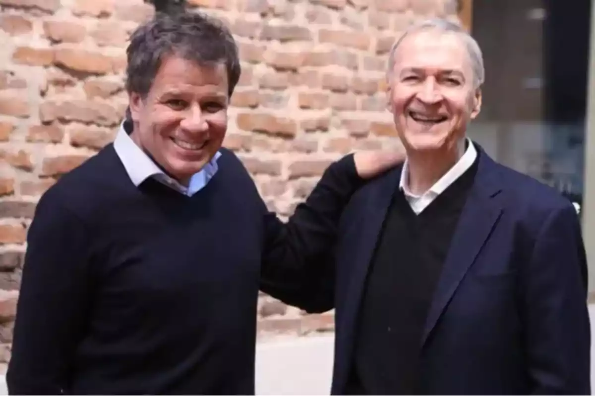 Dos hombres sonrientes posan juntos frente a una pared de ladrillos
