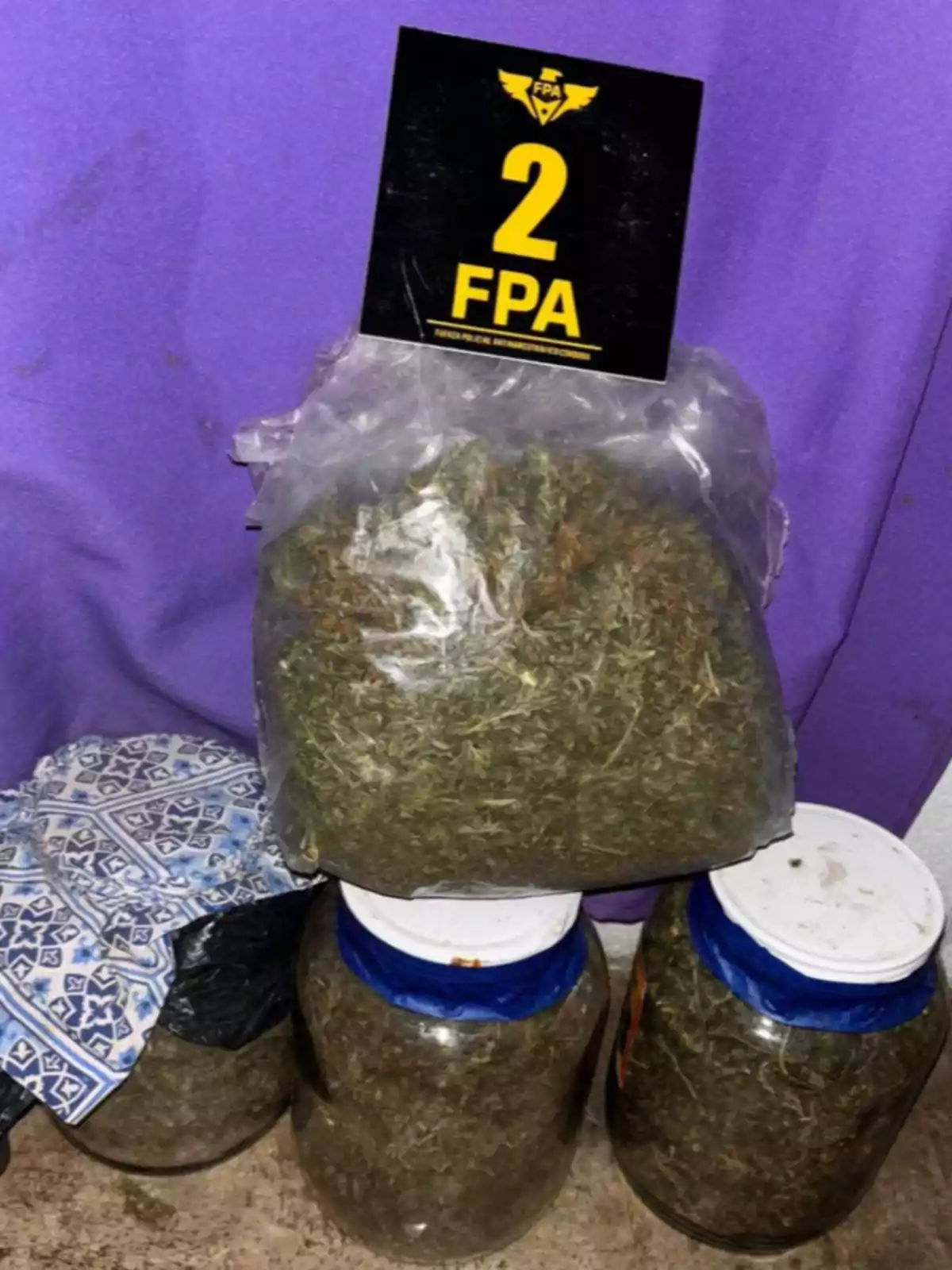 Bolsa y frascos grandes llenos de hierba seca junto a un cartel negro y amarillo con el número 2 y las siglas FPA sobre un fondo violeta