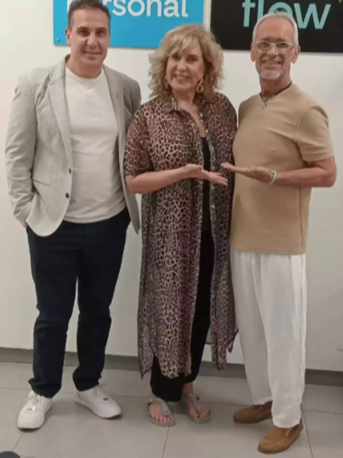 Three people pose smiling in front of a white wall with blue and black posters; the woman in the center is wearing an animal print dress, and the men on either side are dressed in casual clothes. Three people pose smiling in front of a white wall with blue and black posters; the woman in the center is wearing an animal print dress, and the men on either side are dressed in casual clothes.
