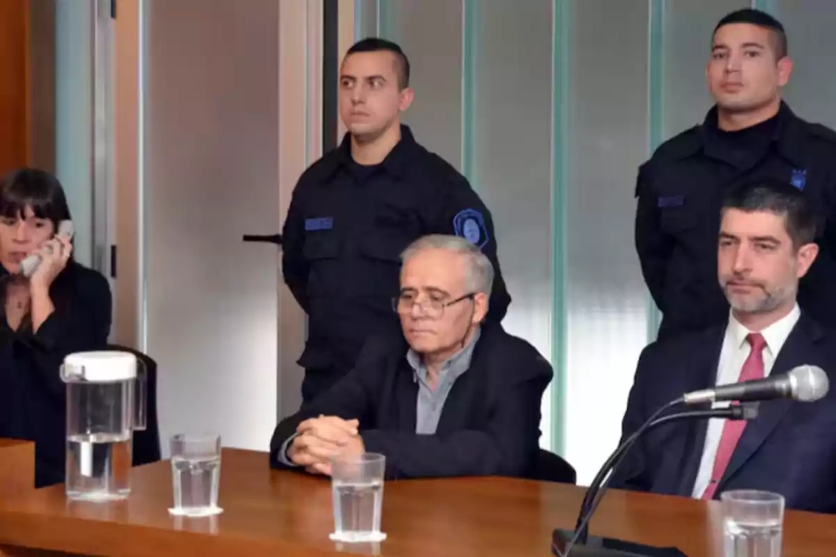 Un hombre mayor con gafas está sentado en una sala de audiencias junto a otras personas, dos policías de pie al fondo y una mujer hablando por teléfono a la izquierda.