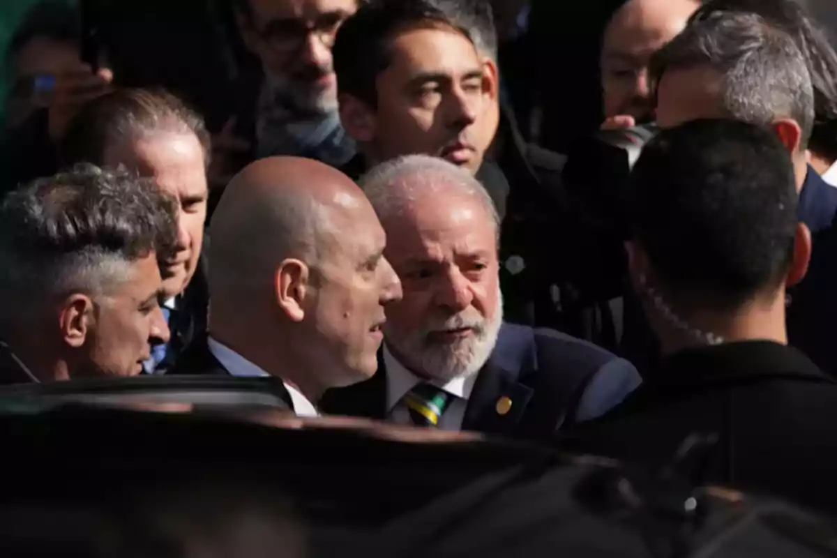 Un grupo de hombres de traje se encuentra reunido en un evento al aire libre, algunos de ellos conversan de cerca mientras otros observan alrededor Un grupo de hombres de traje se encuentra reunido en un evento al aire libre, algunos de ellos conversan de cerca mientras otros observan alrededor