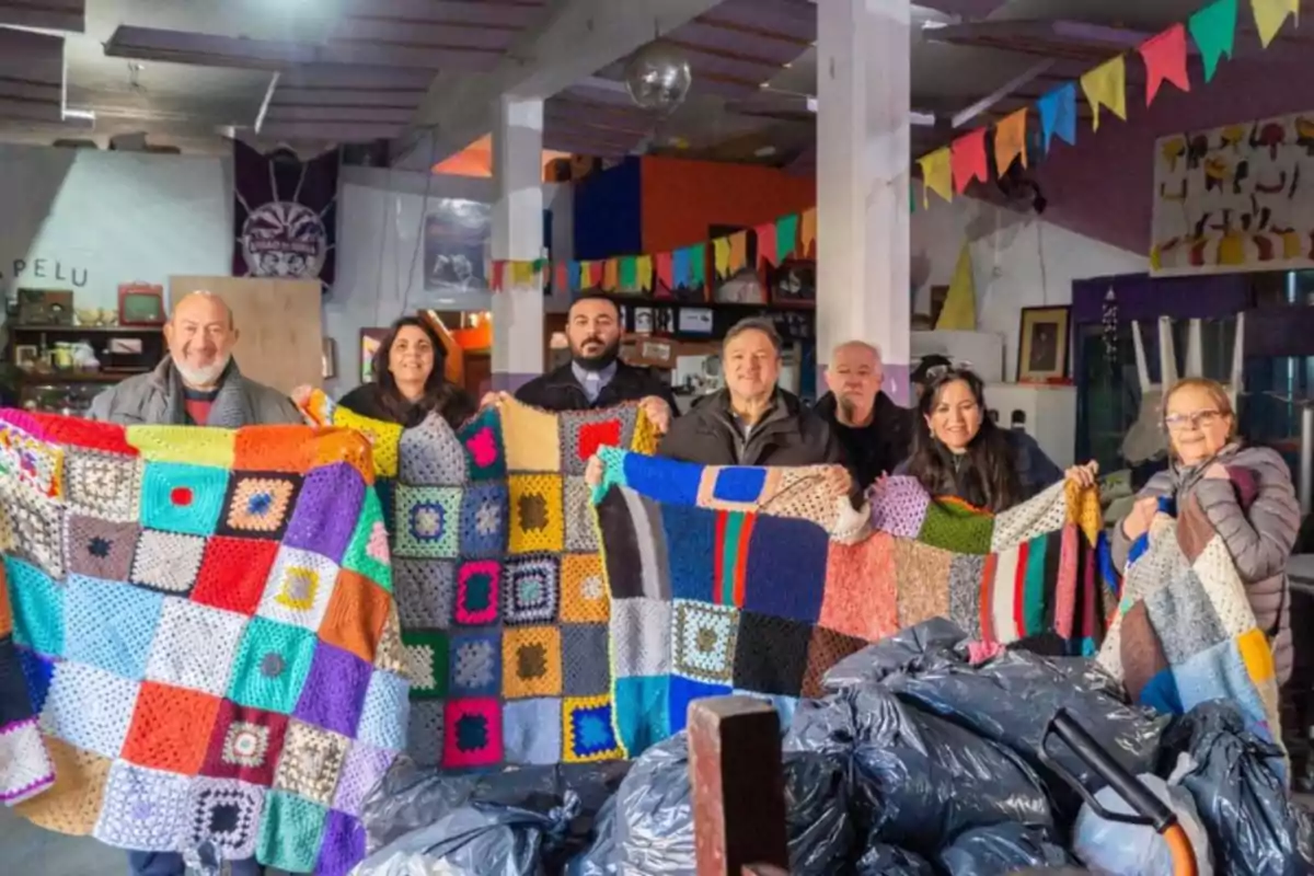 A group of people smiles while holding colorful woven blankets inside a venue decorated with pennants, in front of several black plastic bags.