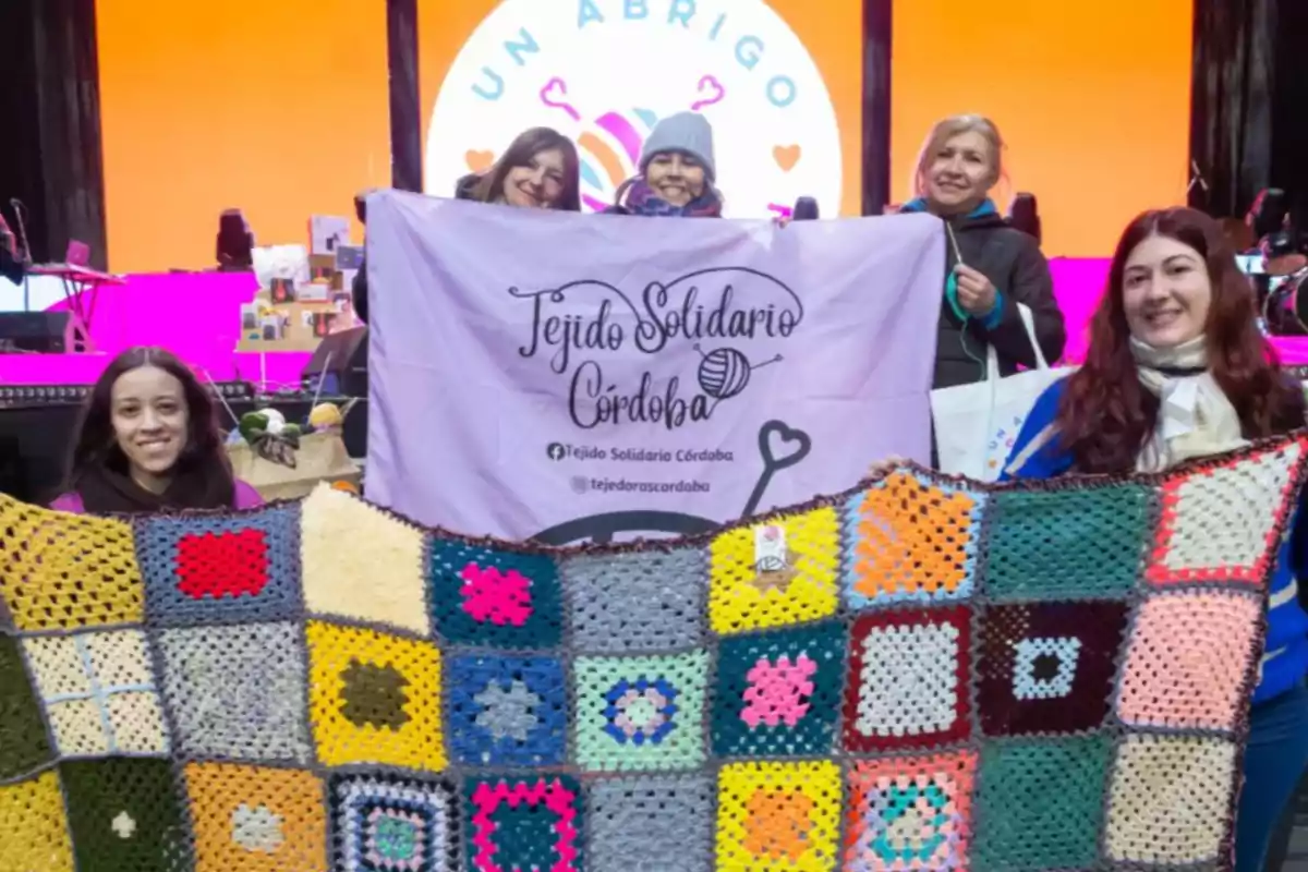 Five women smile while holding a colorful woven blanket and a sign that says Five women smile while holding a colorful woven blanket and a sign that says