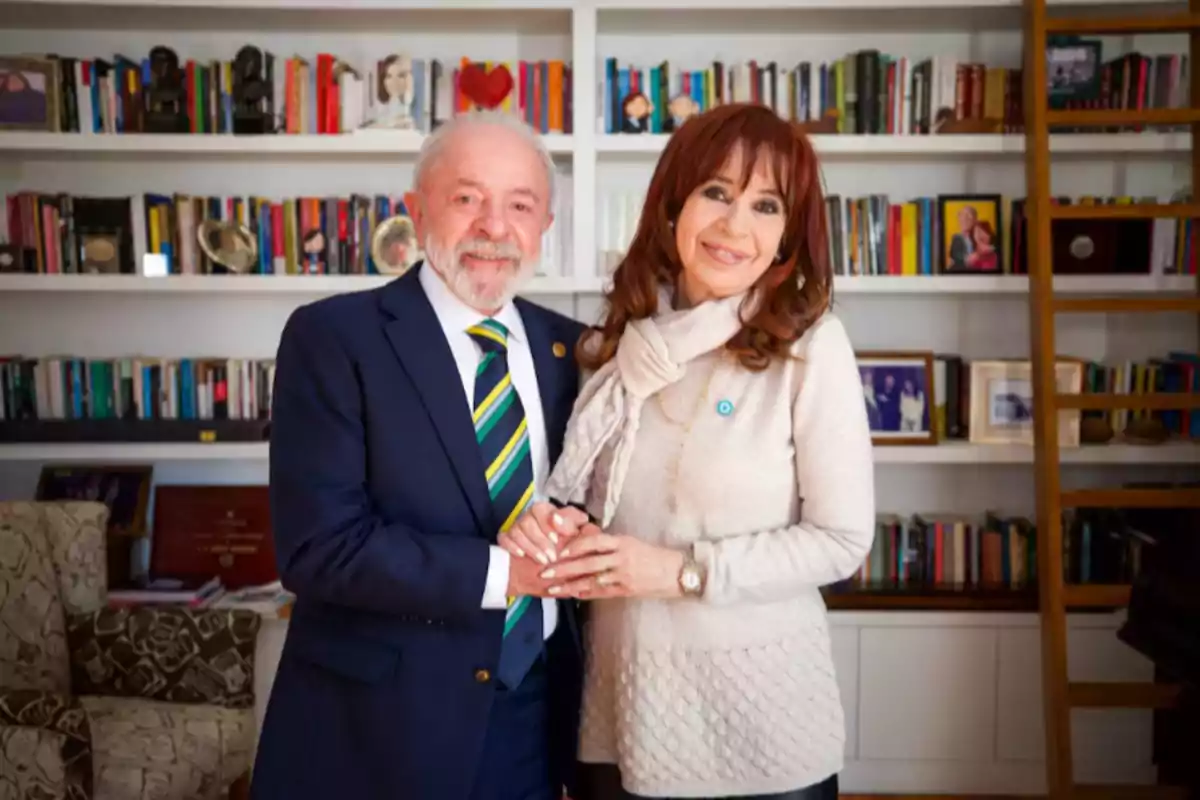 Un hombre de traje y una mujer con suéter claro posan sonrientes tomados de las manos frente a una estantería llena de libros Un hombre de traje y una mujer con suéter claro posan sonrientes tomados de las manos frente a una estantería llena de libros