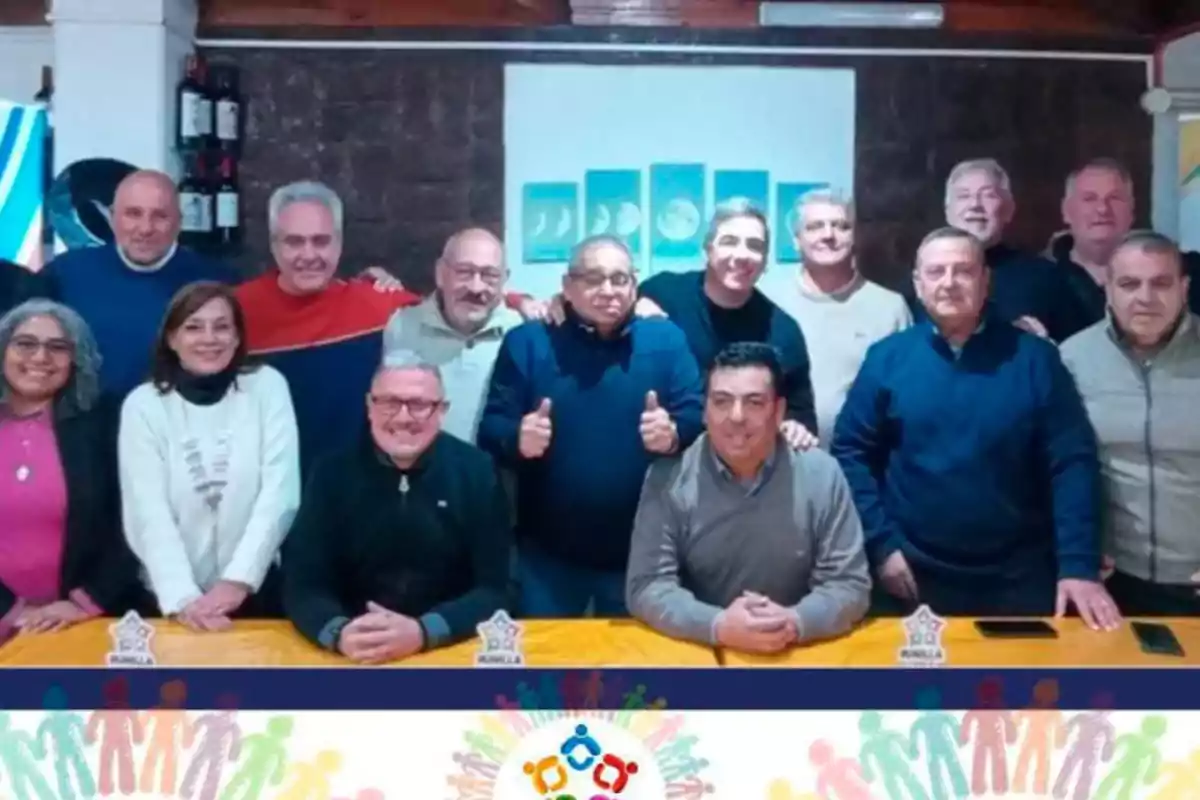 Grupo de personas posando juntas alrededor de una mesa amarilla en un ambiente interior, con una decoración colorida y un cartel con figuras humanas en la parte inferior