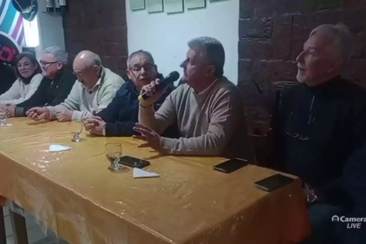 Siete personas sentadas en una mesa cubierta con mantel amarillo, una de ellas sostiene un micrófono mientras las demás escuchan atentamente Siete personas sentadas en una mesa cubierta con mantel amarillo, una de ellas sostiene un micrófono mientras las demás escuchan atentamente