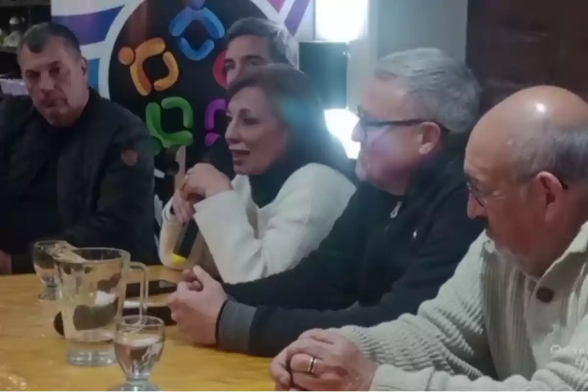 Cinco personas sentadas alrededor de una mesa durante una reunión, con vasos de agua y una jarra sobre la mesa, mientras una mujer sostiene un micrófono y habla Cinco personas sentadas alrededor de una mesa durante una reunión, con vasos de agua y una jarra sobre la mesa, mientras una mujer sostiene un micrófono y habla