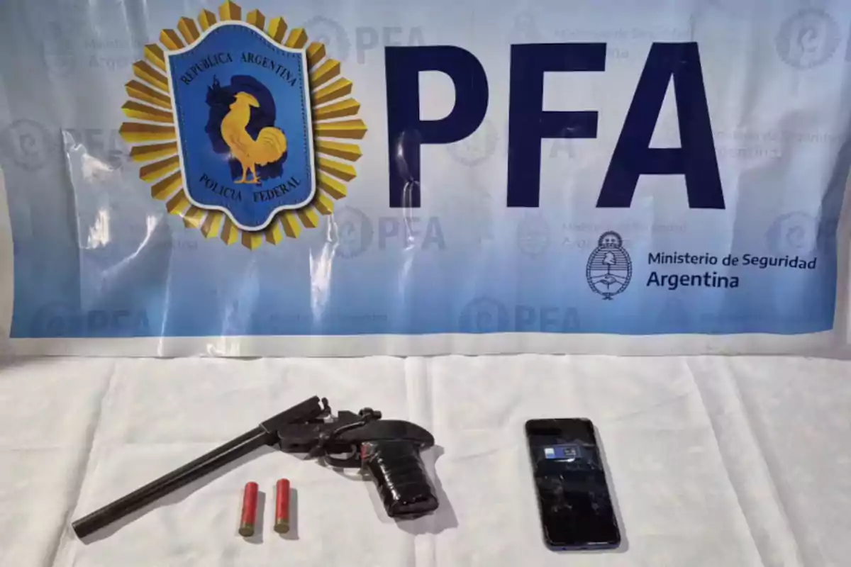 Sobre una mesa blanca hay una pistola, dos cartuchos y un teléfono móvil, al fondo se ve un cartel de la Policía Federal Argentina. Sobre una mesa blanca hay una pistola, dos cartuchos y un teléfono móvil, al fondo se ve un cartel de la Policía Federal Argentina.