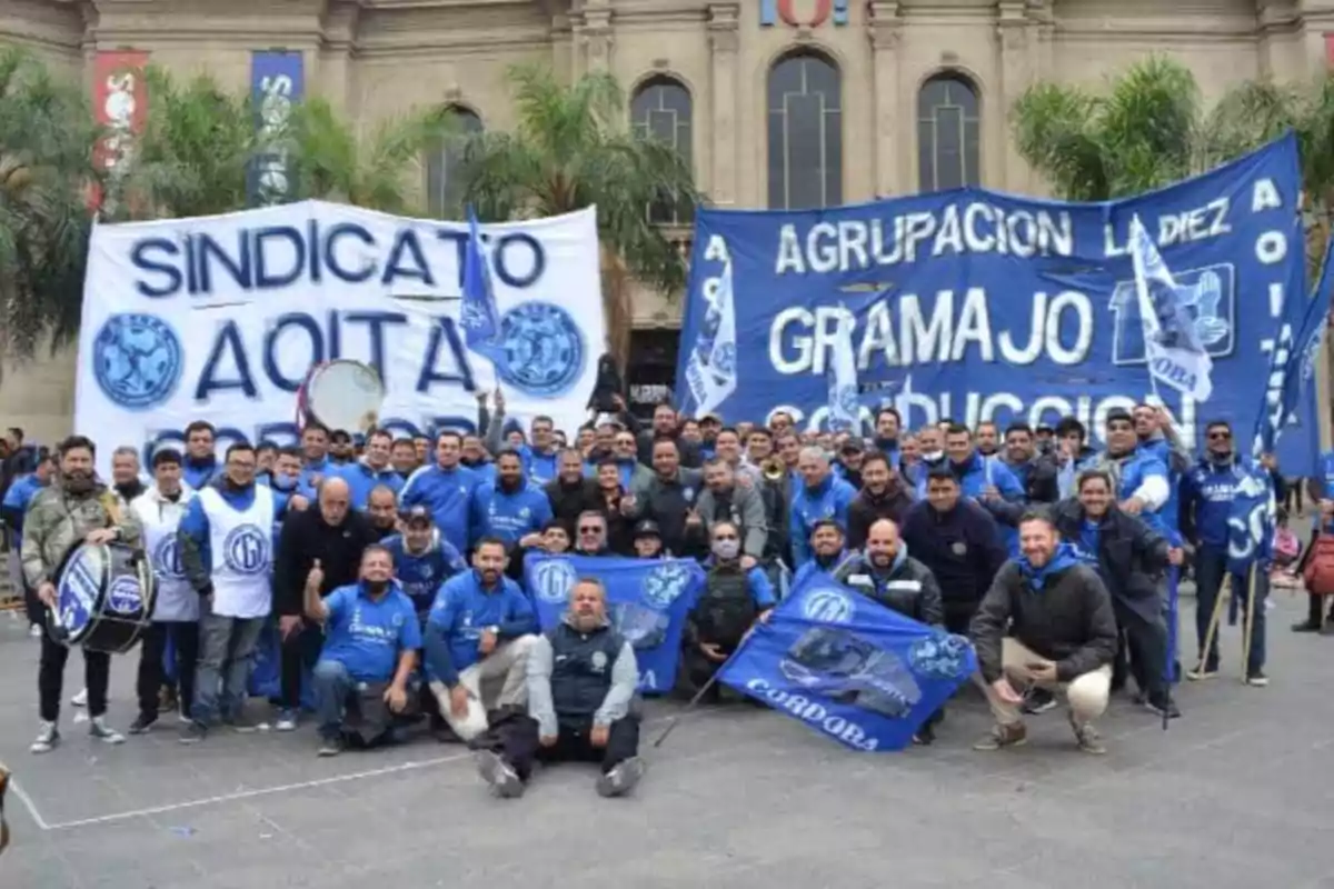 A large group of people poses outdoors with flags and blue jerseys in front of an old building, displaying union and group banners. A large group of people poses outdoors with flags and blue jerseys in front of an old building, displaying union and group banners.