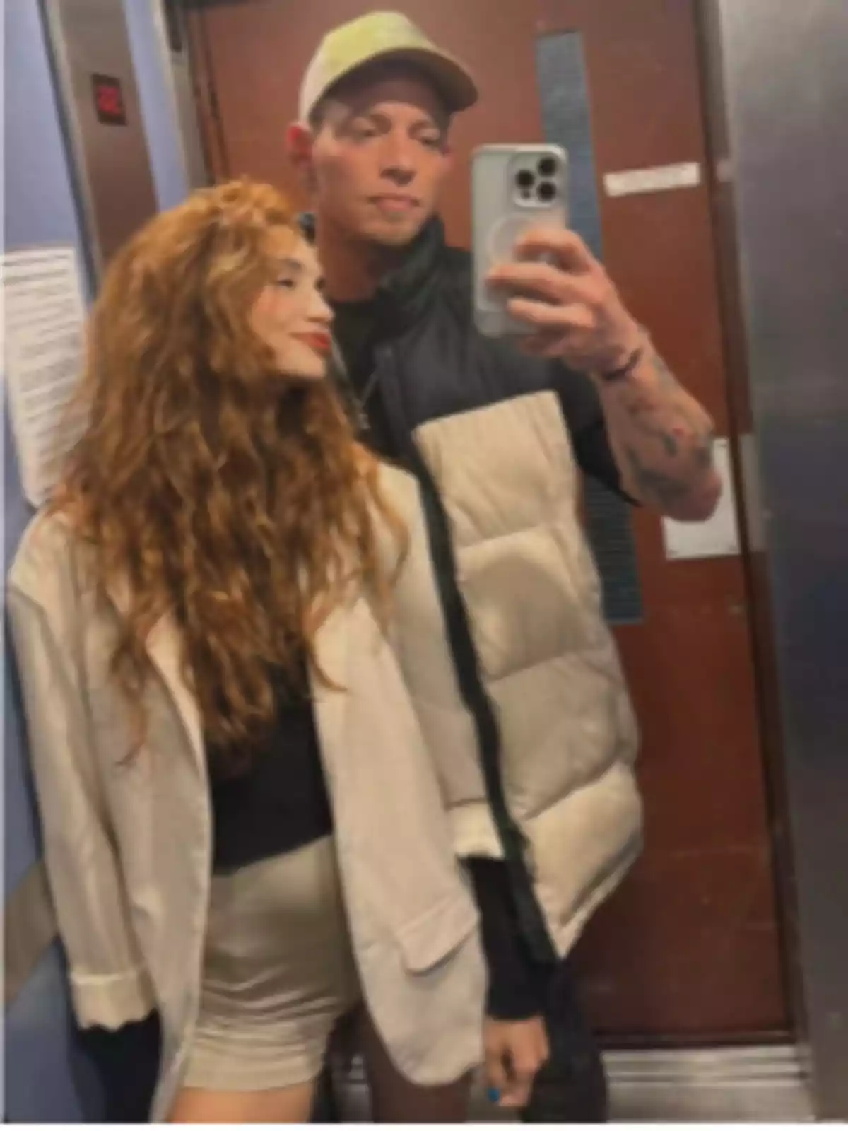 Two people pose for a selfie in the mirror of an elevator, one of them holding the phone while the other smiles and looks at it. Two people pose for a selfie in the mirror of an elevator, one of them holding the phone while the other smiles and looks at it.