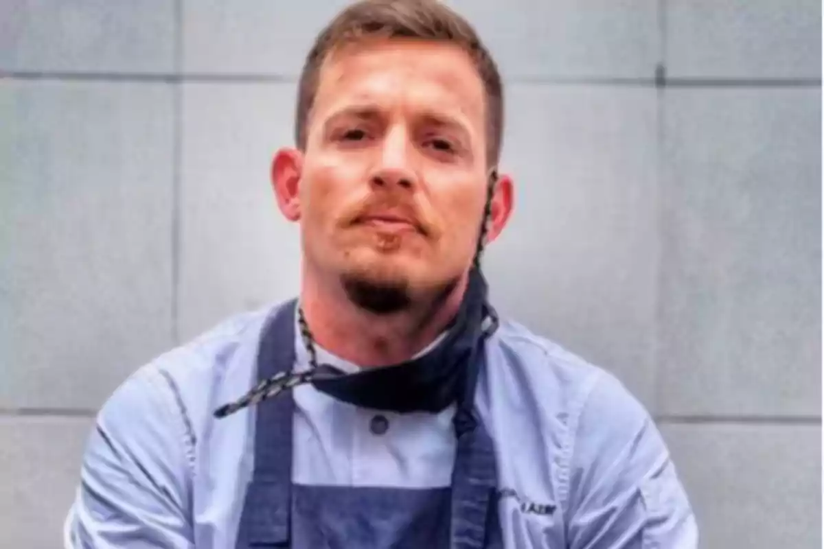 Man with a short beard and short hair wears a light jersey and a dark apron in front of a gray wall Man with a short beard and short hair wears a light jersey and a dark apron in front of a gray wall
