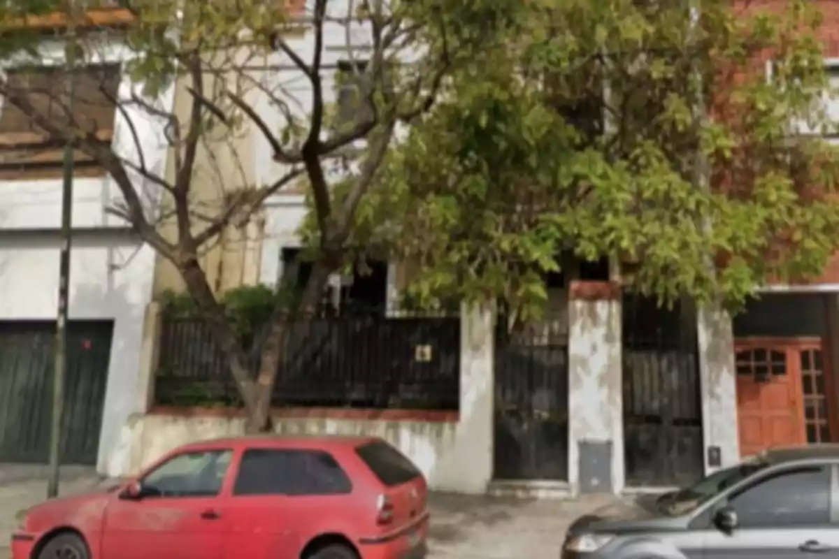 Fachada de una casa con rejas negras y árboles al frente, un auto rojo y otro gris estacionados en la calle Fachada de una casa con rejas negras y árboles al frente, un auto rojo y otro gris estacionados en la calle