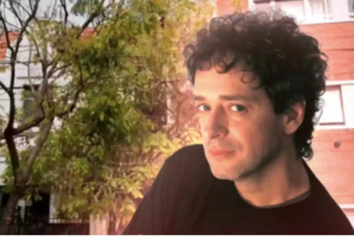 Hombre de cabello rizado y oscuro con camiseta negra posando frente a un fondo de árboles y edificios.