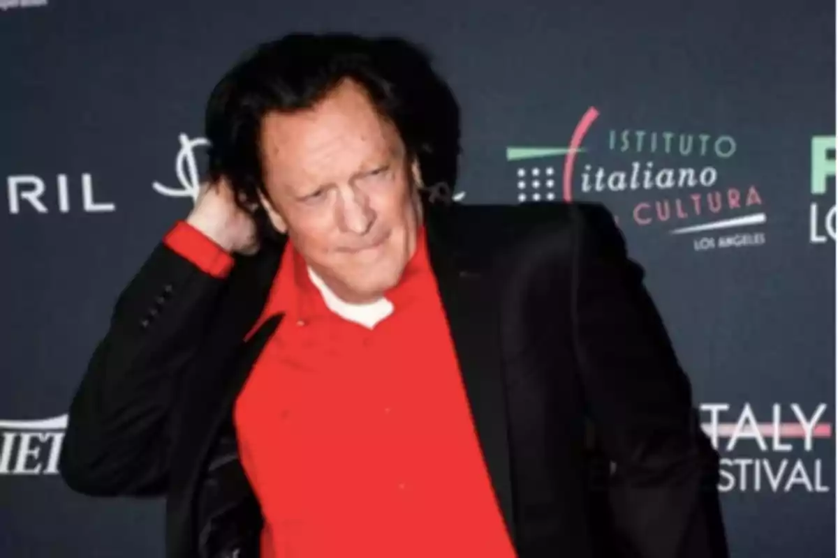 Dark-haired man wearing a red jersey and black jacket posing in front of a backdrop with cultural event logos Dark-haired man wearing a red jersey and black jacket posing in front of a backdrop with cultural event logos