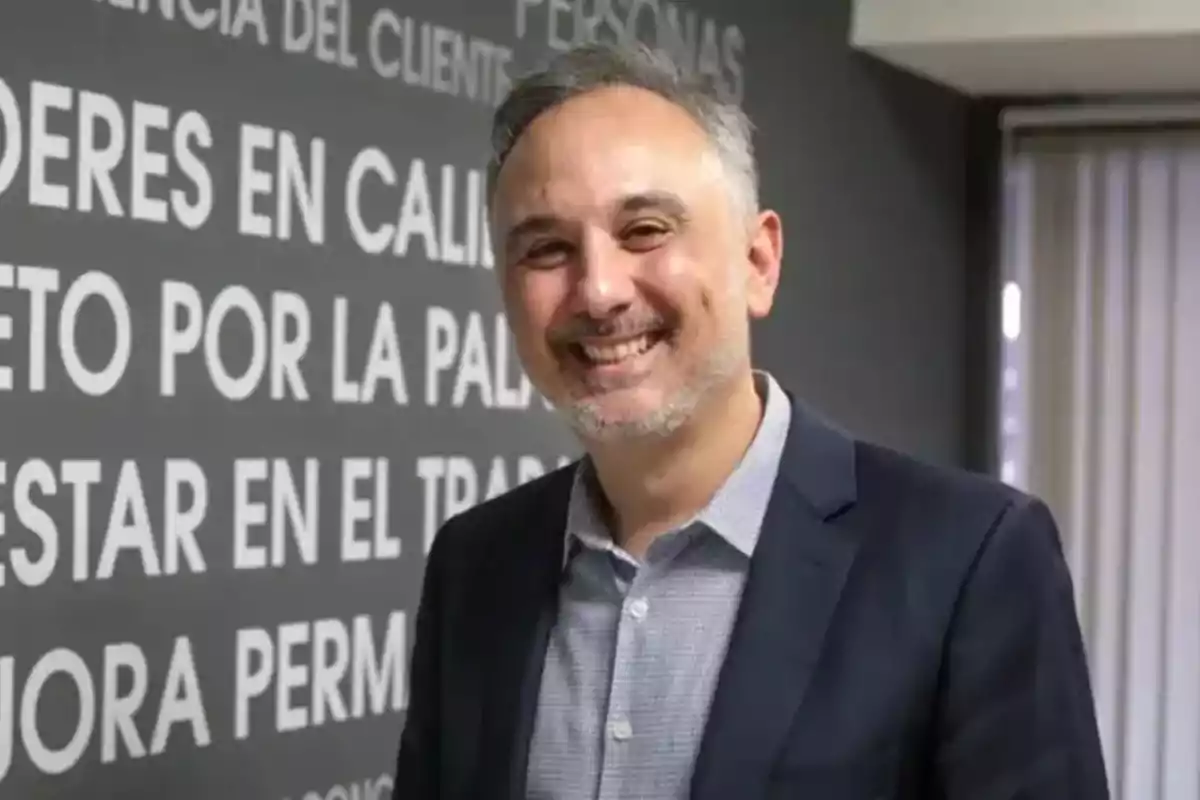 Gray-haired man with a short beard smiling, wearing a blue jacket and a light jersey, standing in front of a gray wall with white-lettered text