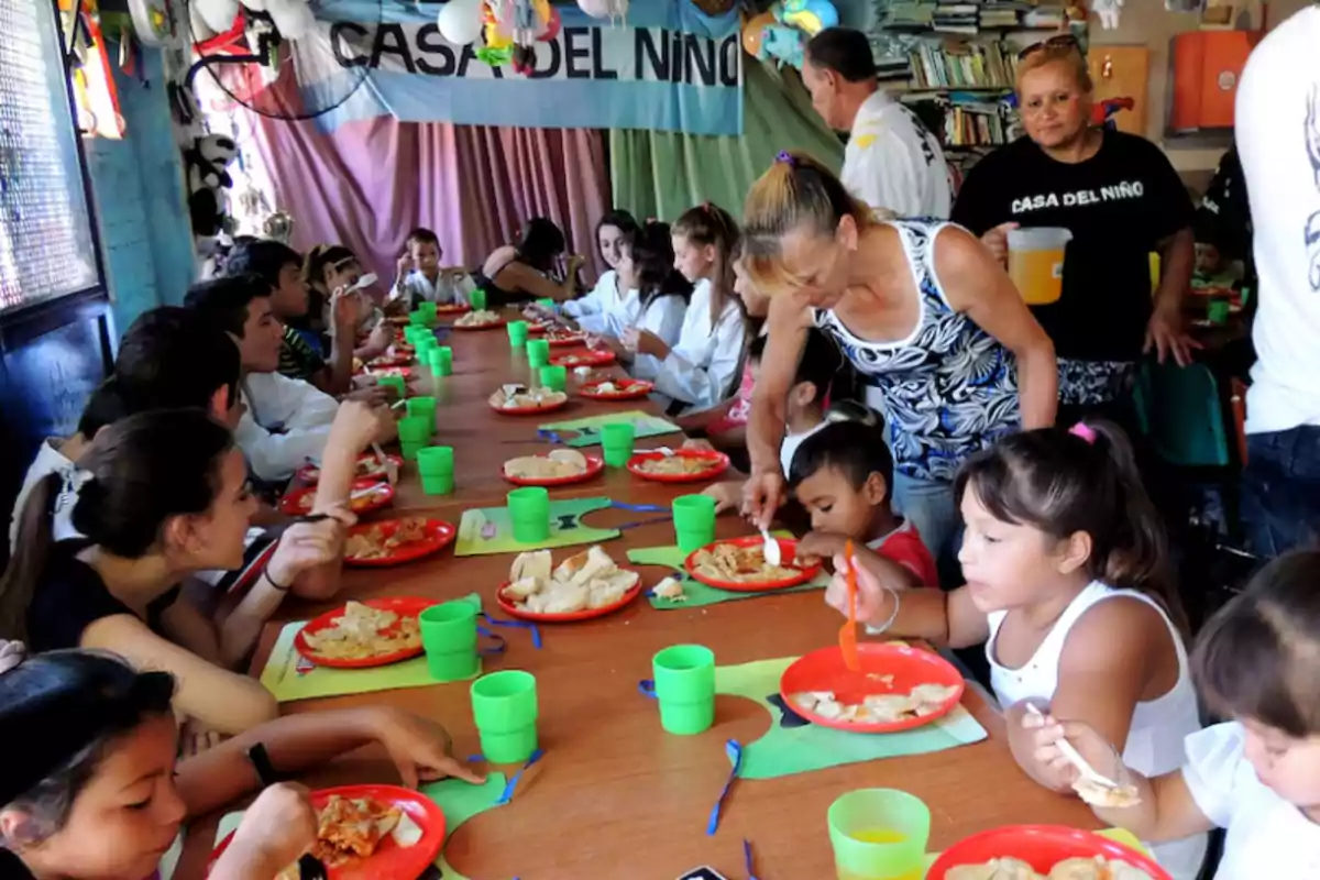 Un grupo de niños y adultos comparte una comida en una mesa larga dentro de un comedor comunitario decorado con globos y carteles, mientras una mujer ayuda a servir la comida y otra reparte bebida.