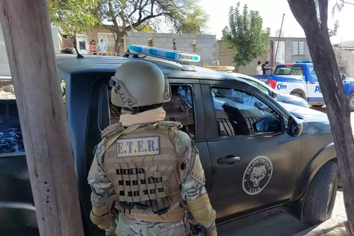 Un agente táctico con uniforme de camuflaje y casco se encuentra junto a una camioneta negra con insignias policiales en una zona urbana donde también hay patrullas y personas al fondo. Un agente táctico con uniforme de camuflaje y casco se encuentra junto a una camioneta negra con insignias policiales en una zona urbana donde también hay patrullas y personas al fondo.